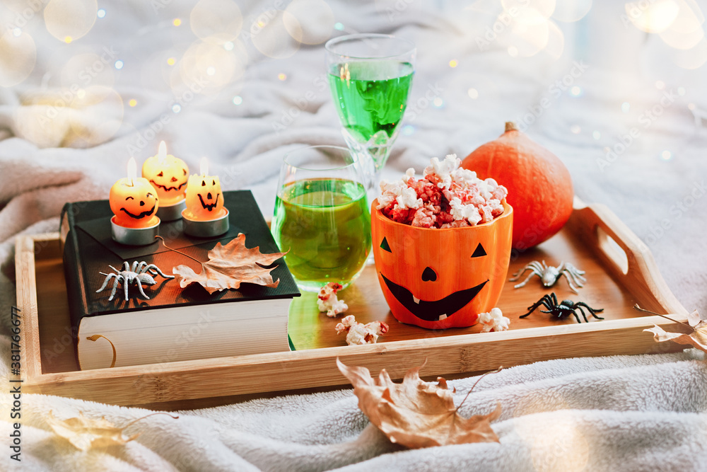 Book, halloween decor, candles, popcorn and drinks on tray on bed, halloween reading concept with bokeh background