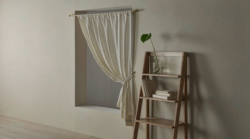Plain undamaged cream wall in a rental flat with a sheer curtain on a brass tension rod inside the window recess and a freestanding oak ladder shelf