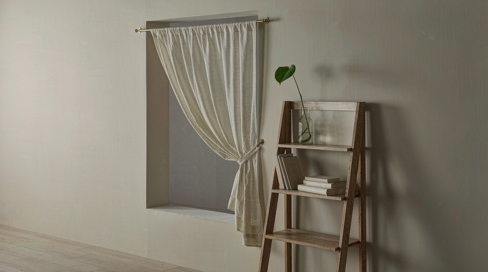 Plain undamaged cream wall in a rental flat with a sheer curtain on a brass tension rod inside the window recess and a freestanding oak ladder shelf