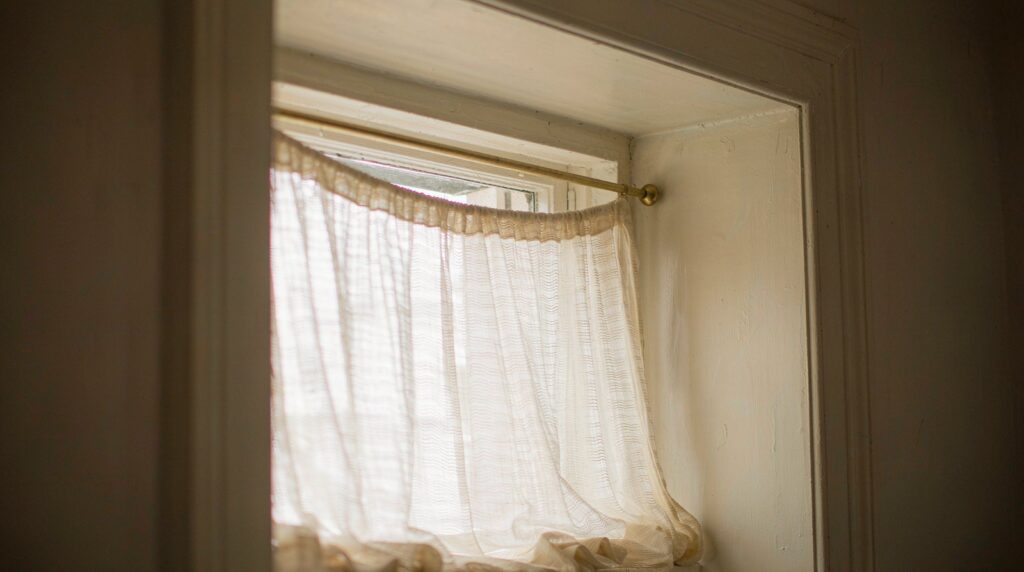 Slim brass tension rod pressed into both sides of a window recess holding a sheer curtain, with no screws or brackets visible