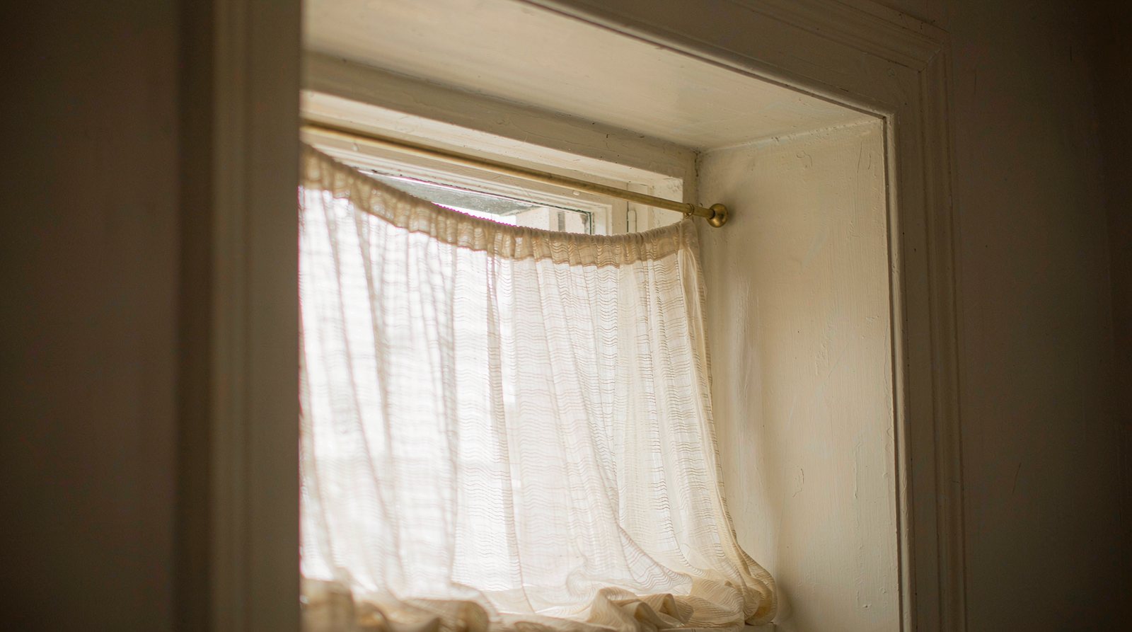 Slim brass tension rod pressed into both sides of a window recess holding a sheer curtain, with no screws or brackets visible